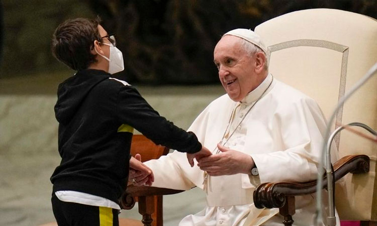 A imagem mostra um momento de carinho e acolhimento entre o Papa Francisco e uma criança. O Papa está sentado em uma grande cadeira com braços de madeira, vestindo sua tradicional batina branca, solidéu (o pequeno chapéu branco na cabeça) e um crucifixo pendurado ao pescoço. Ele sorri calorosamente enquanto segura as mãos de um menino que está em pé à sua frente. O menino usa óculos, máscara branca de proteção e veste um agasalho preto com listras amarelas nas laterais e calça combinando. Ele se inclina levemente para frente, demonstrando atenção e afeto. O cenário sugere que o encontro acontece dentro de uma igreja ou salão religioso, com luz suave e ambiente solene. A imagem transmite uma forte sensação de empatia, acolhimento e respeito.